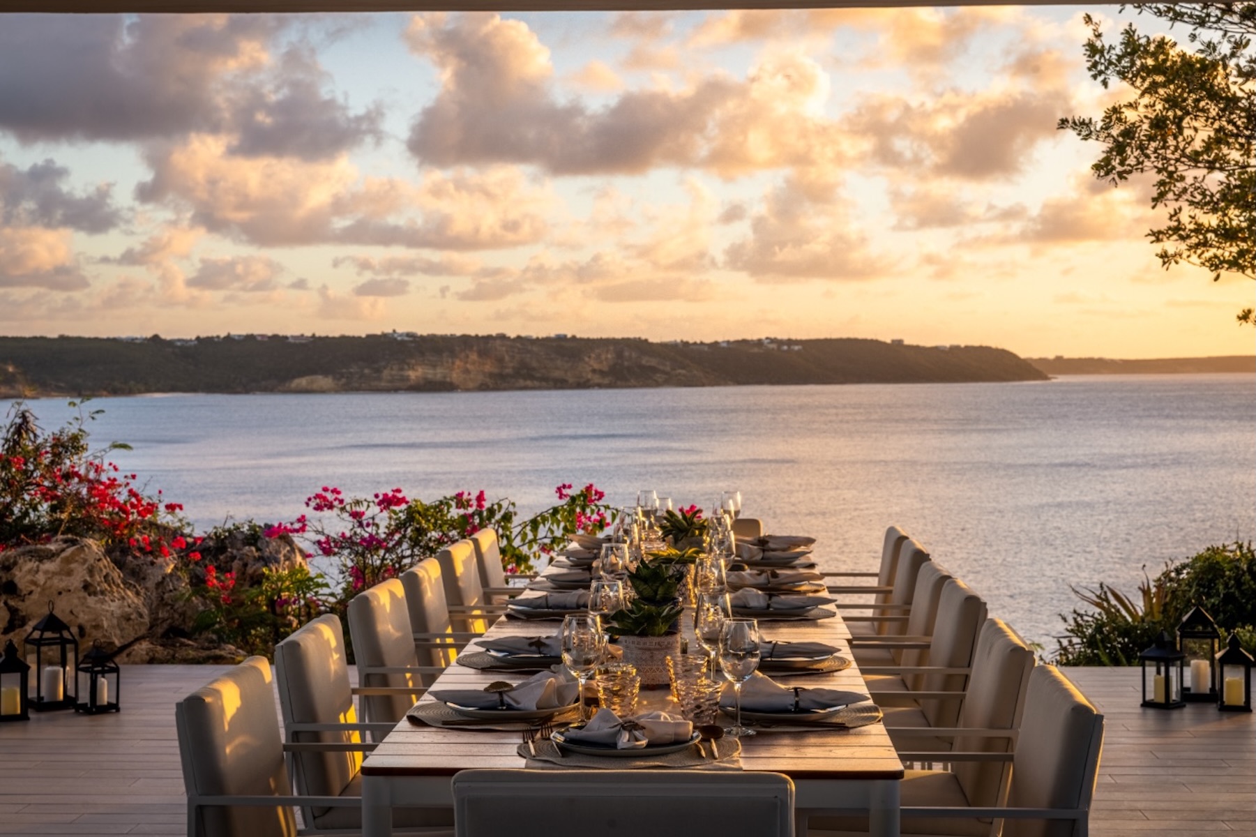 Elegant dining setup on a terrace overlooking the ocean at sunset, adorned with flowers and lanterns.