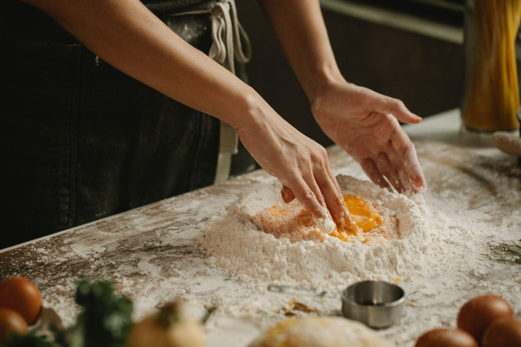 Hands mixing flour and egg yolks on a wooden surface, with fresh ingredients and kitchen tools in the background.
