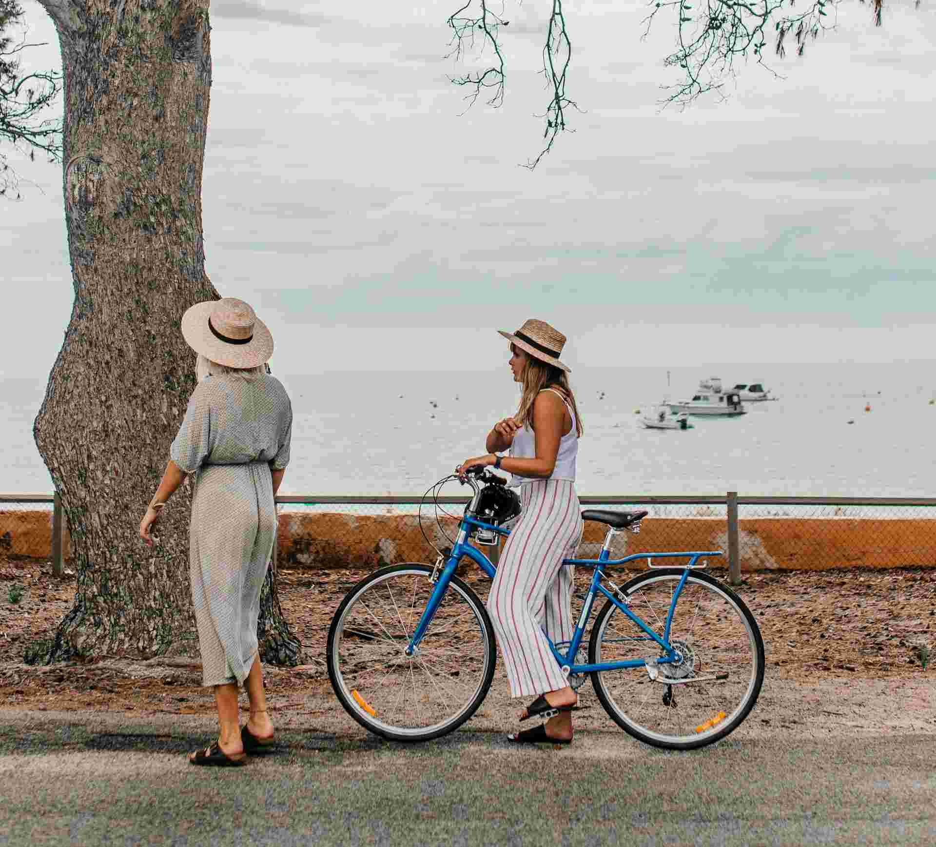 Two women in hats stroll along a seaside path, one holding a bike, enjoying the coastal scenery.